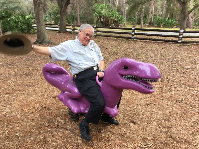 Dan Hopkins riding a purple dinosaur at a park and waving his hat in the hair.