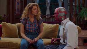 Wanda sits on yellow couch in tears as Vision kneels beside her in his work clothes.
