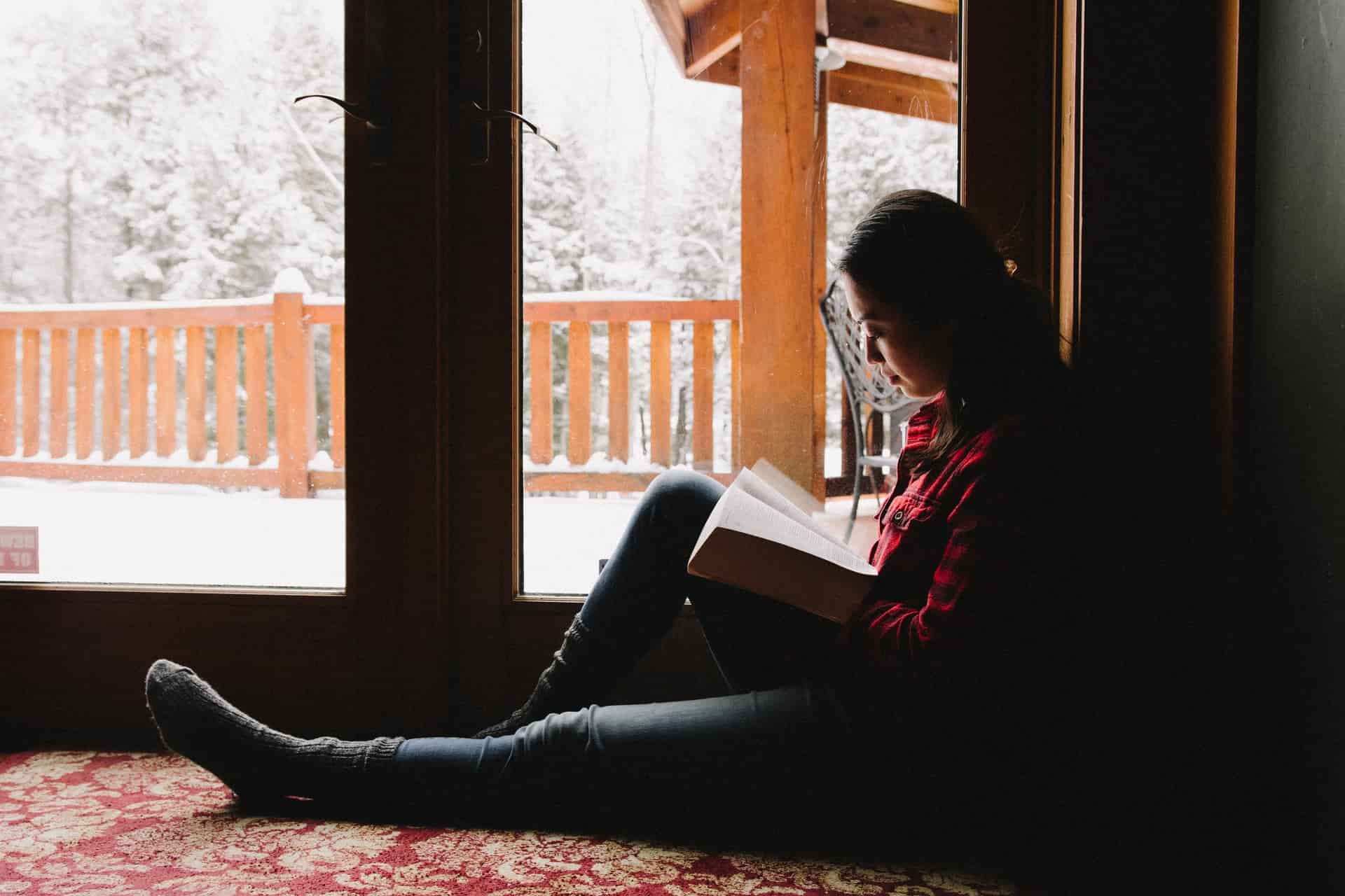 Woman sitting by a lighted window, reading the Bible on a snowy day.